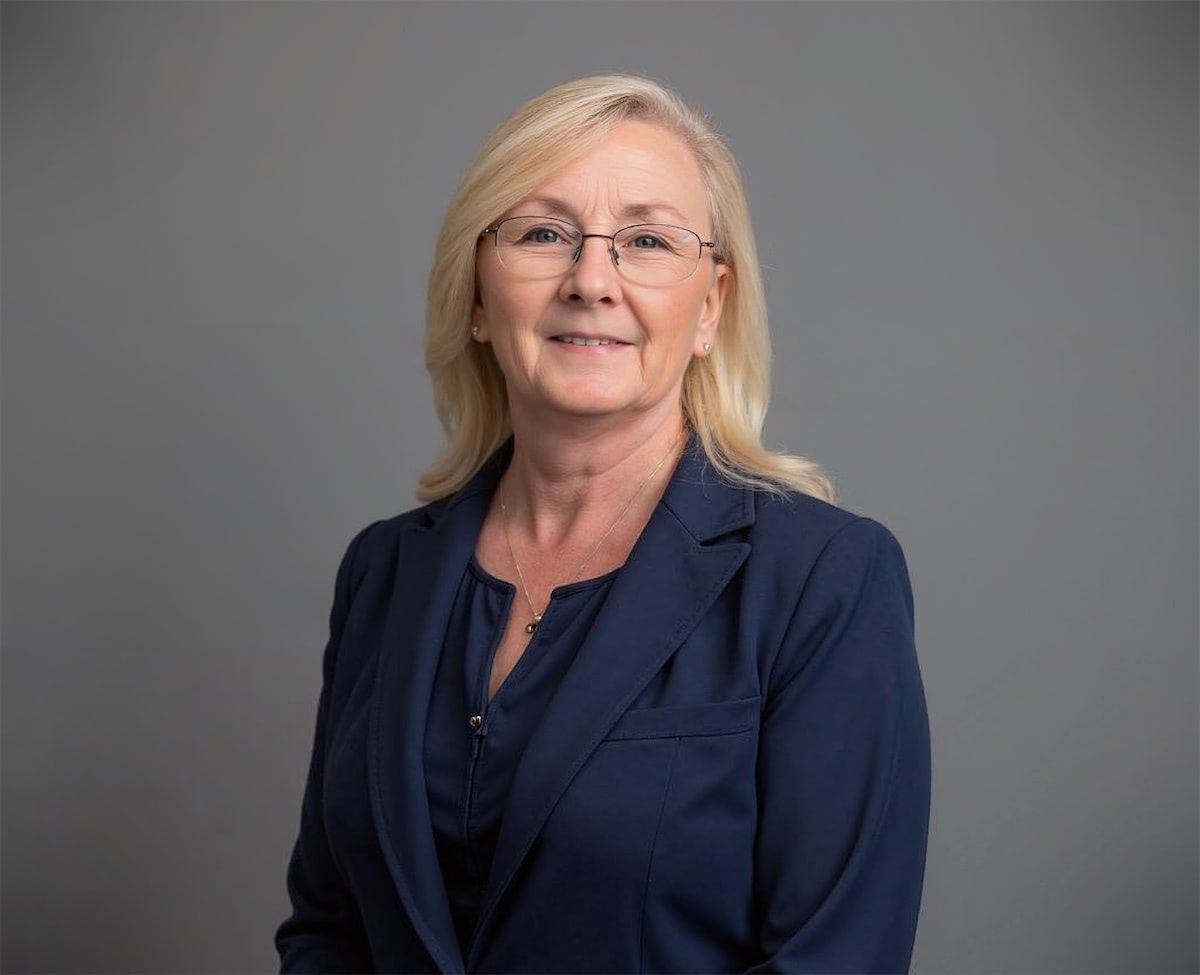 A professional headshot of a person with blonde hair and glasses, wearing a dark blue blazer against a gray background.