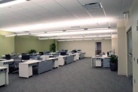 A modern, well-lit open-plan office with rows of desks, gray carpet, and light green walls.