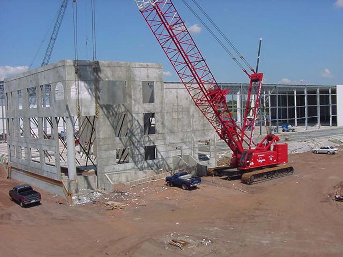 A large red crane lifts a concrete wall panel at a construction site with other buildings visible in the background.