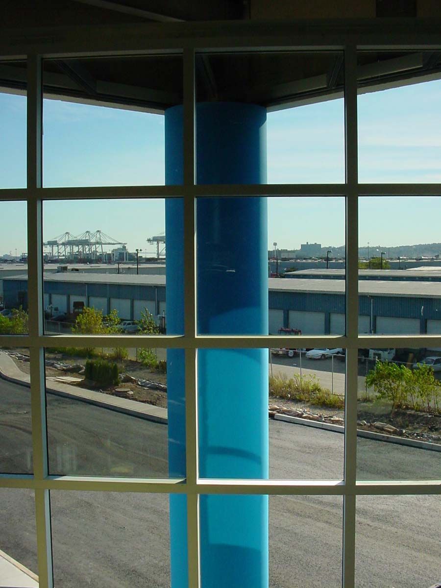 A view through a white grid window showing a large blue pillar and an industrial landscape with cranes and warehouses.
