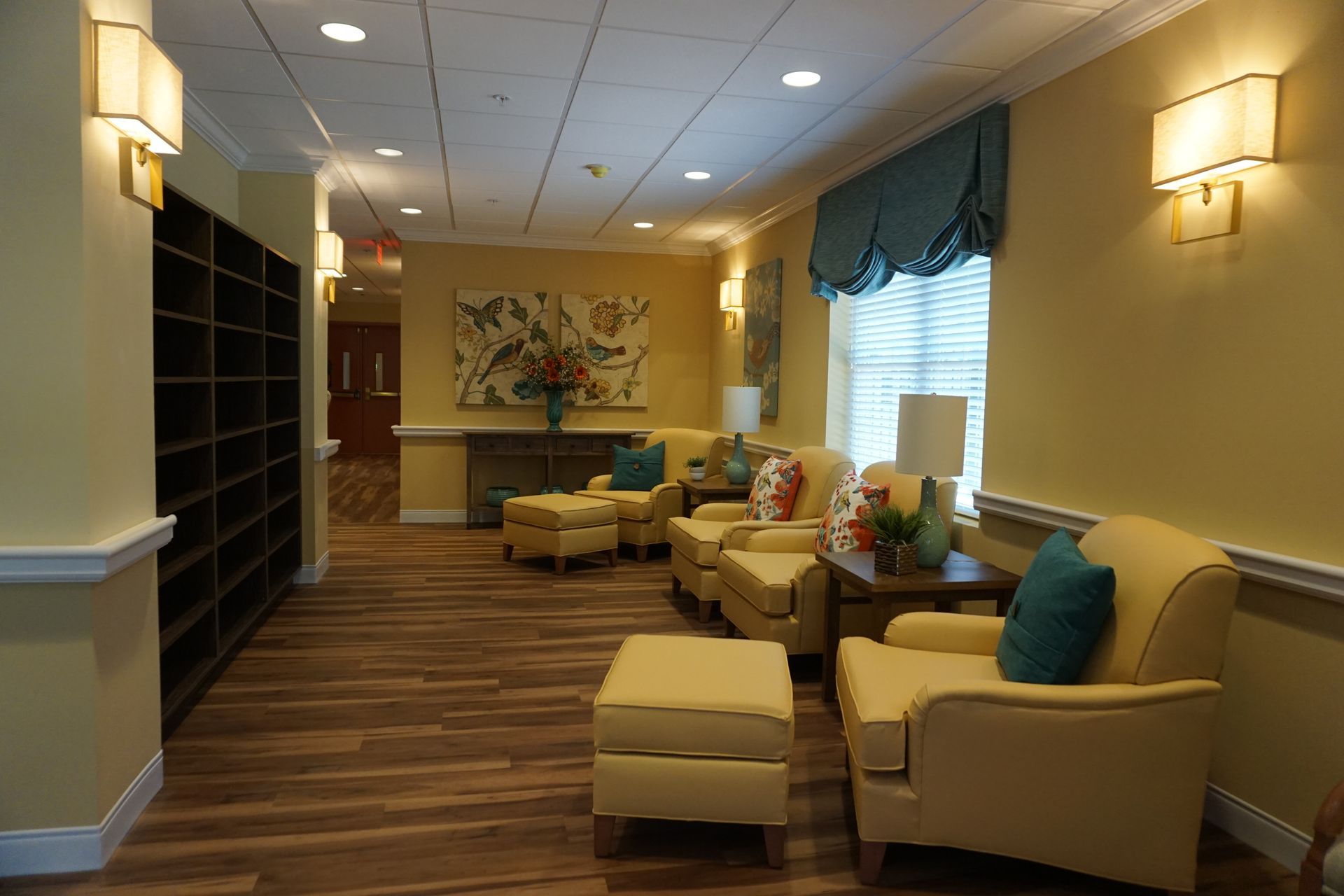 A bright, cozy waiting area with yellow armchairs, ottomans, wood floors, wall sconces, and artwork in a neutral hallway.
