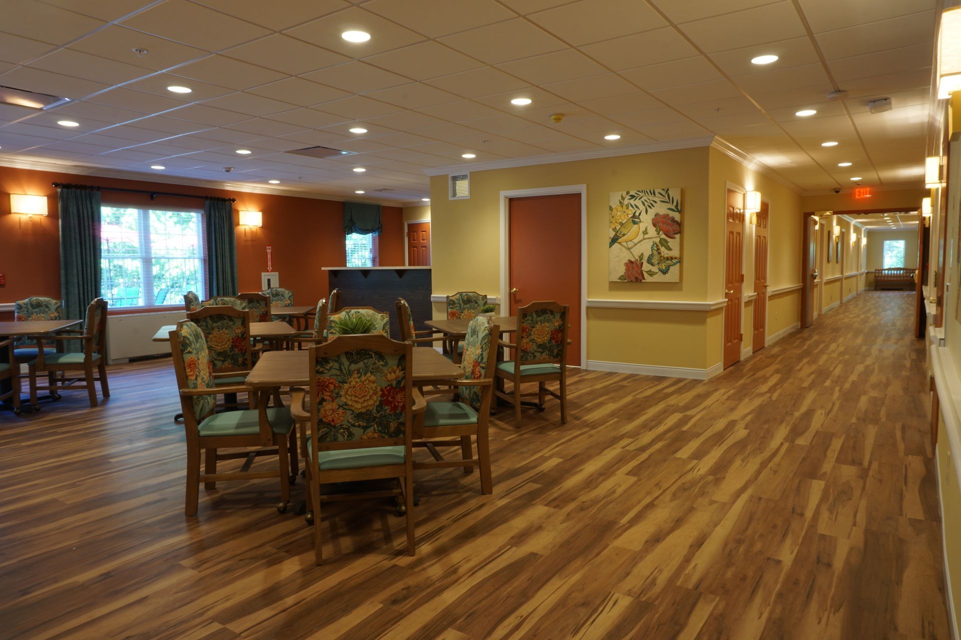 A dining room in a care facility with tables and patterned chairs, leading to a long hallway with hardwood floors.