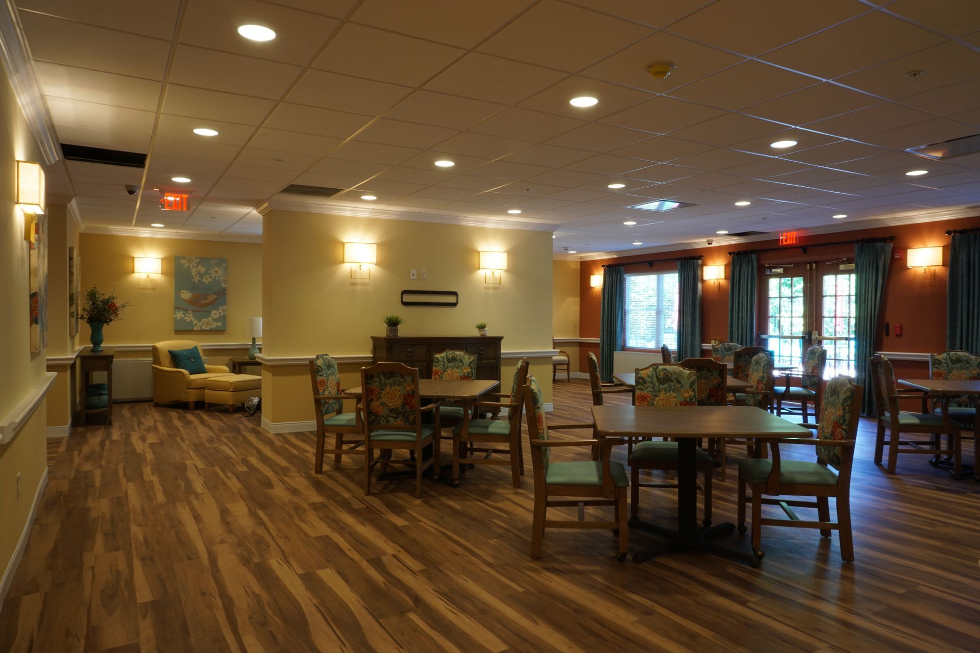 An indoor dining or common area with tables, chairs, wood-look flooring, yellow walls, and warm lighting.