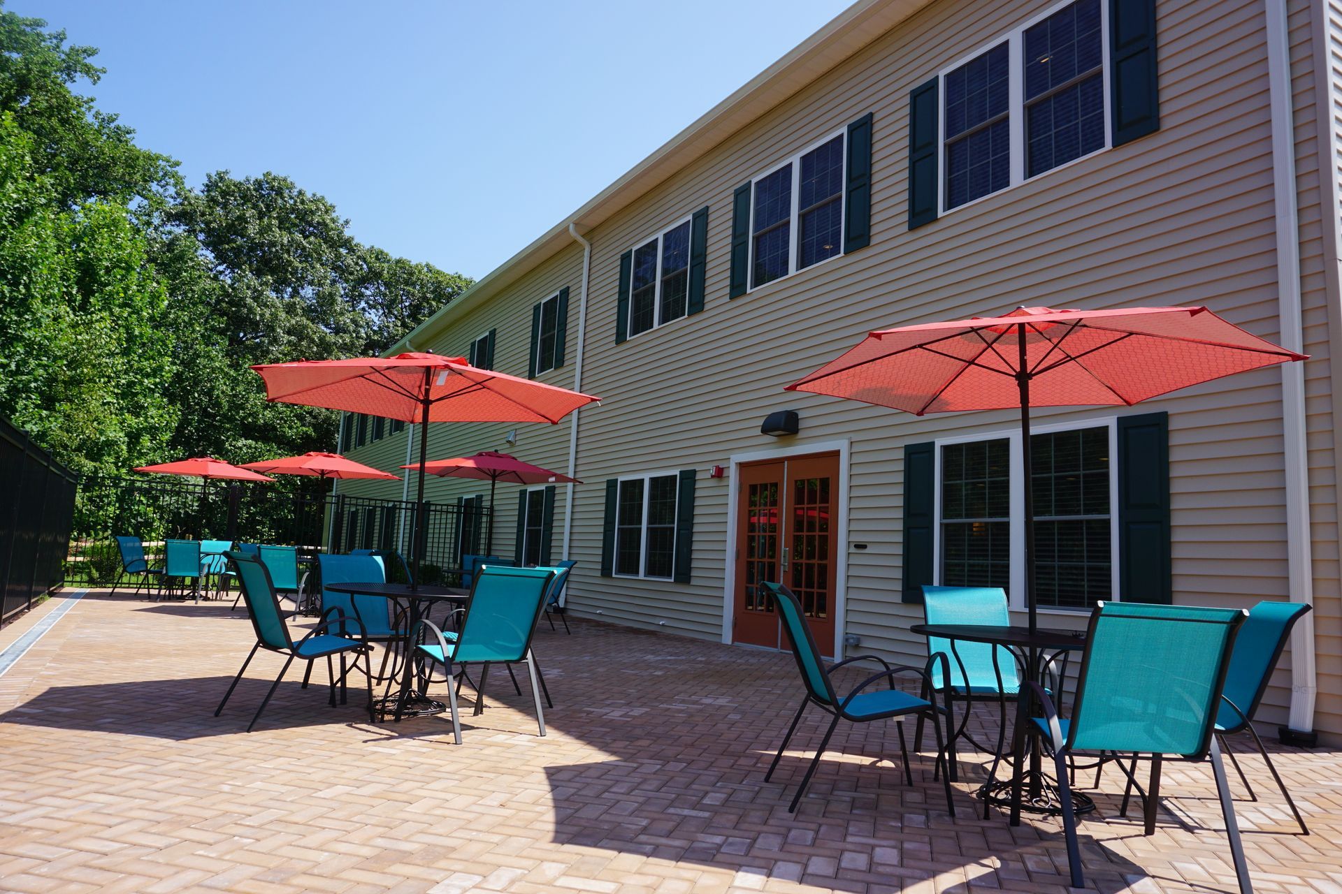 Outdoor patio with several tables, teal chairs, and red umbrellas in front of a two-story beige building with dark shutters.