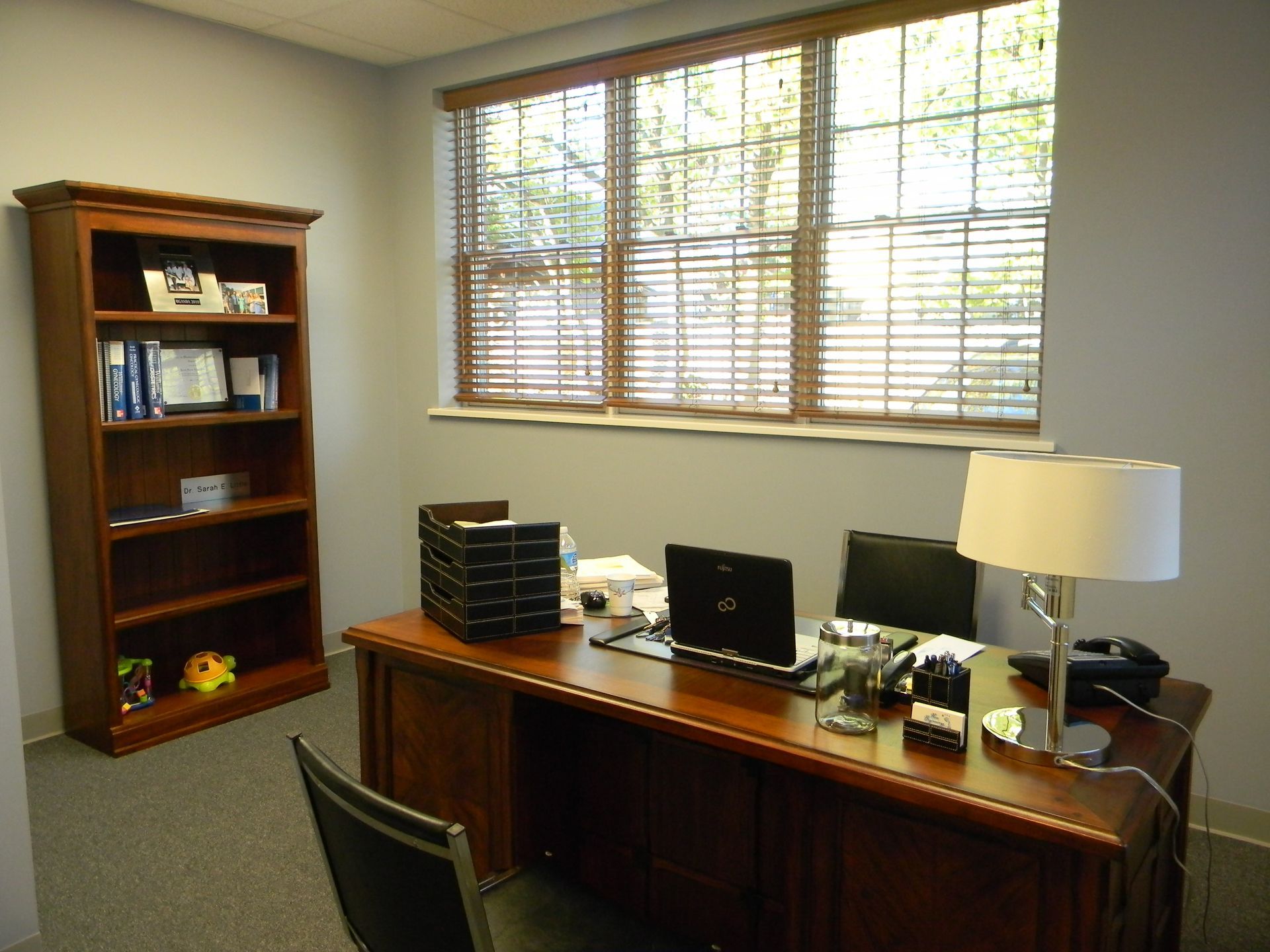 A home or office workspace featuring a wooden desk with a laptop, lamp, and chair next to a wooden bookshelf near a window.
