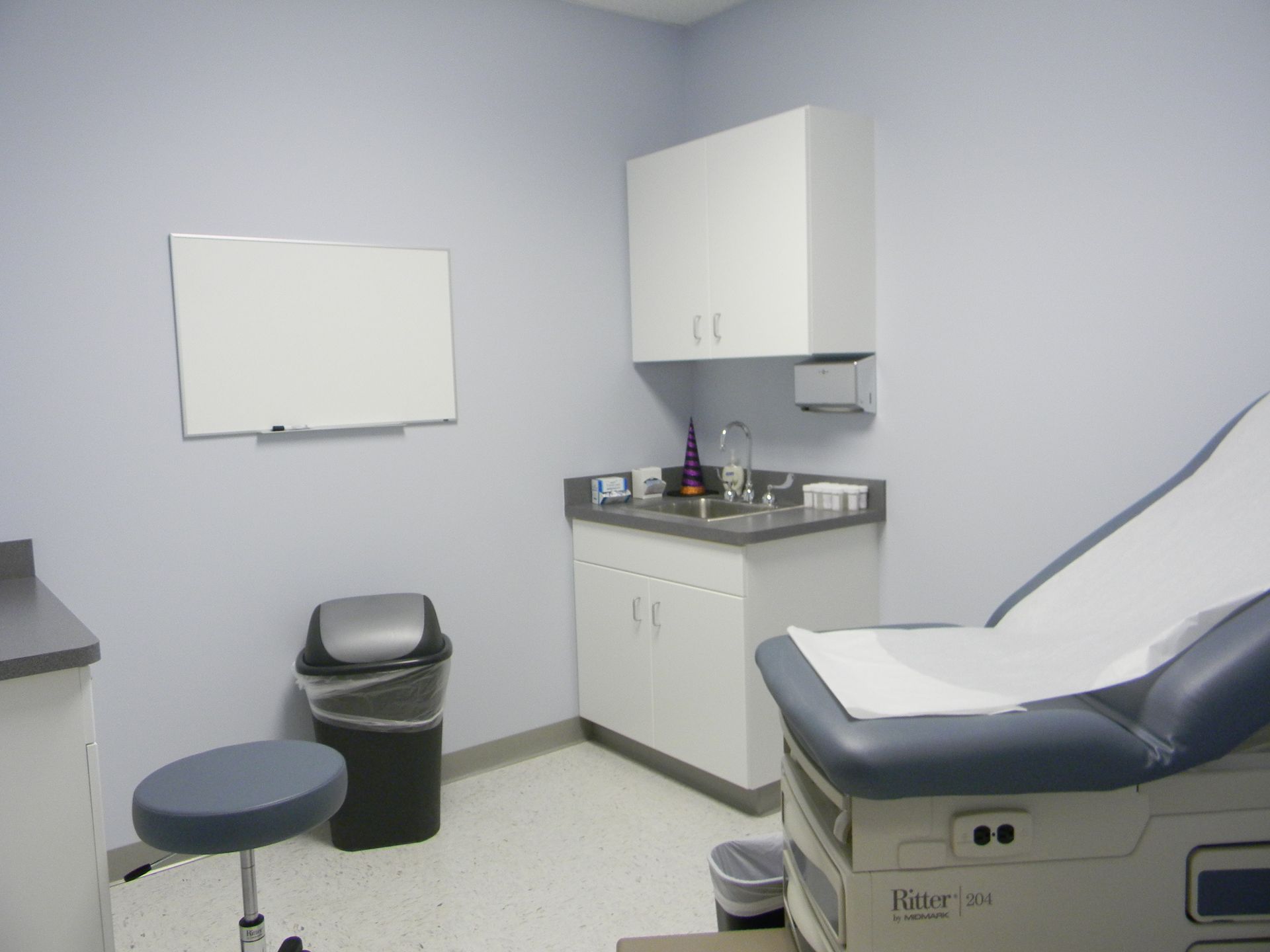 An exam room with blue-grey walls, a medical table, a stool, a sink with cabinets, and a whiteboard.