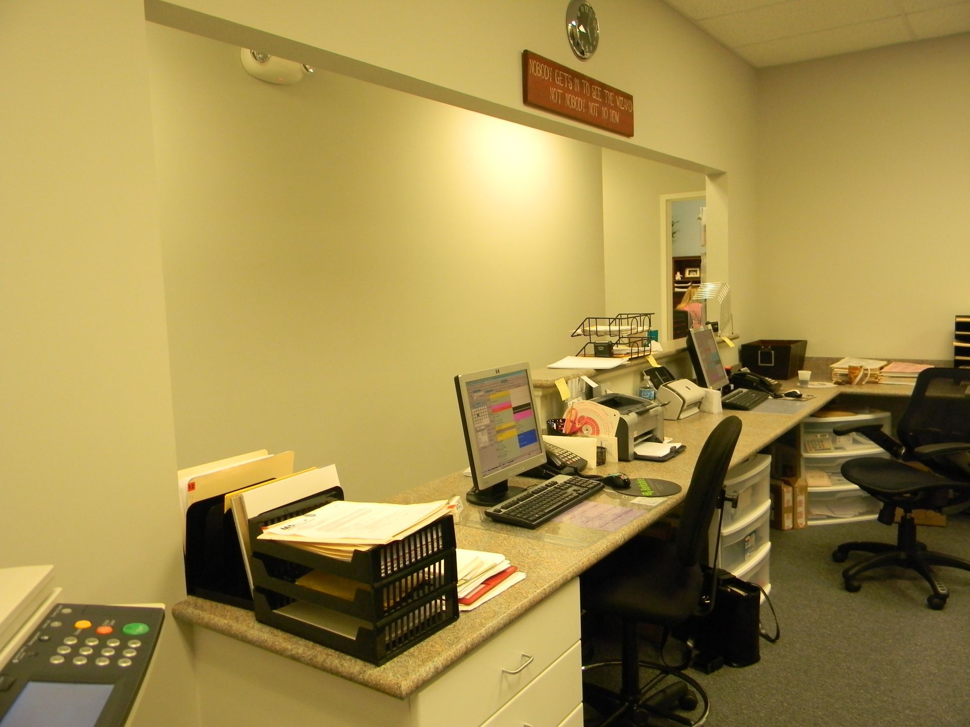 An office reception desk with a computer, black chairs, and stacked papers against a plain wall.