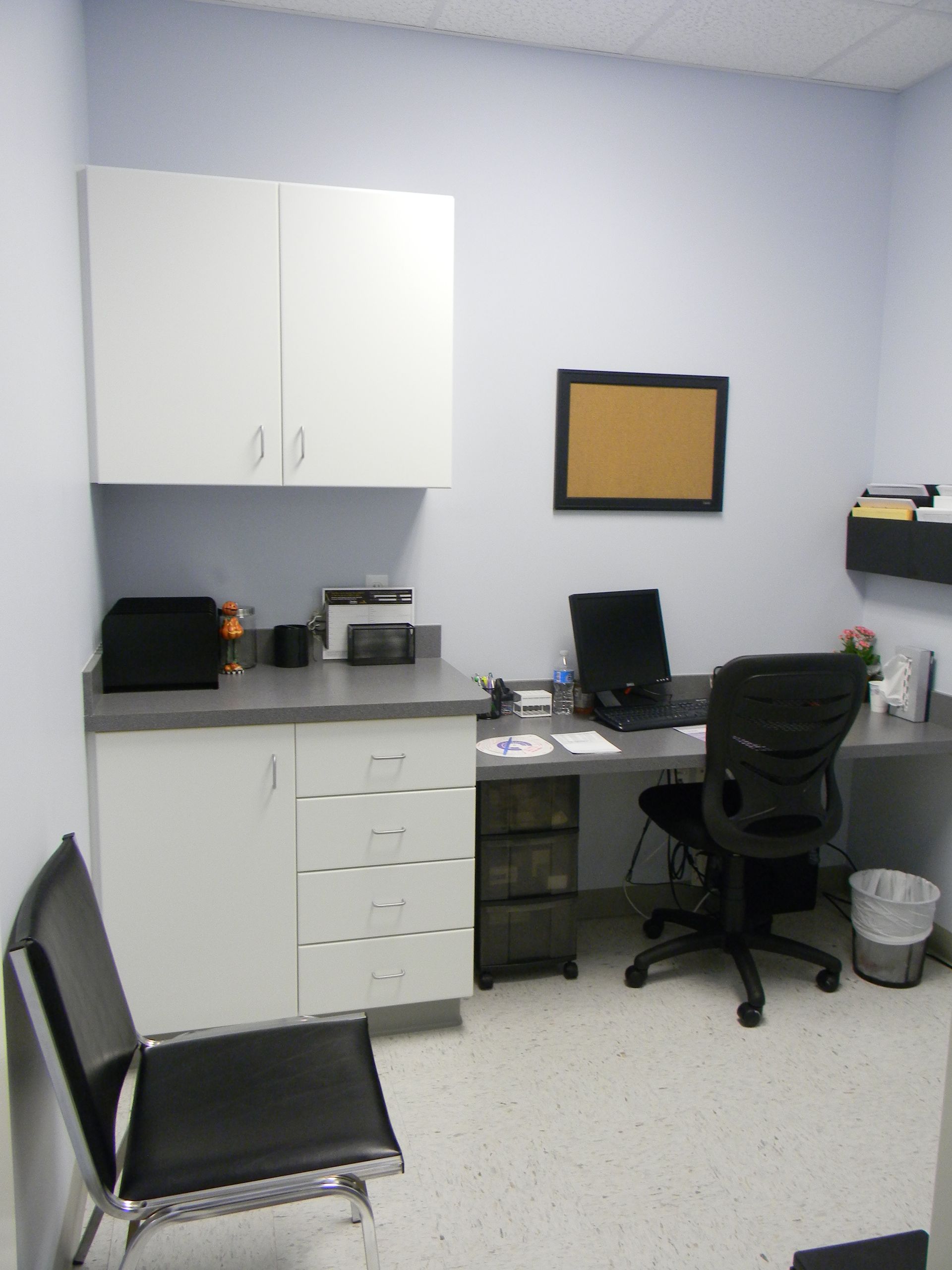 A medical examination room with white cabinets, a desk with a computer, an office chair, and a black guest chair.