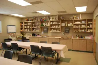 A classroom with a long table covered in a pink cloth, surrounded by chairs, facing shelves filled with ceramic objects.