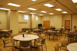A well-lit, spacious community room featuring several round tables and chairs arranged for group gatherings.