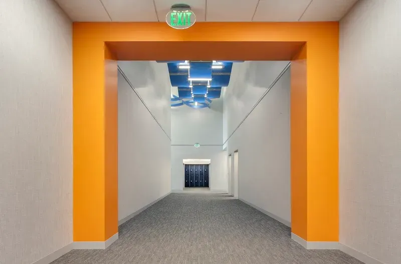A wide, carpeted hallway framed by bright orange walls, featuring a geometric blue ceiling sculpture and distant lockers.