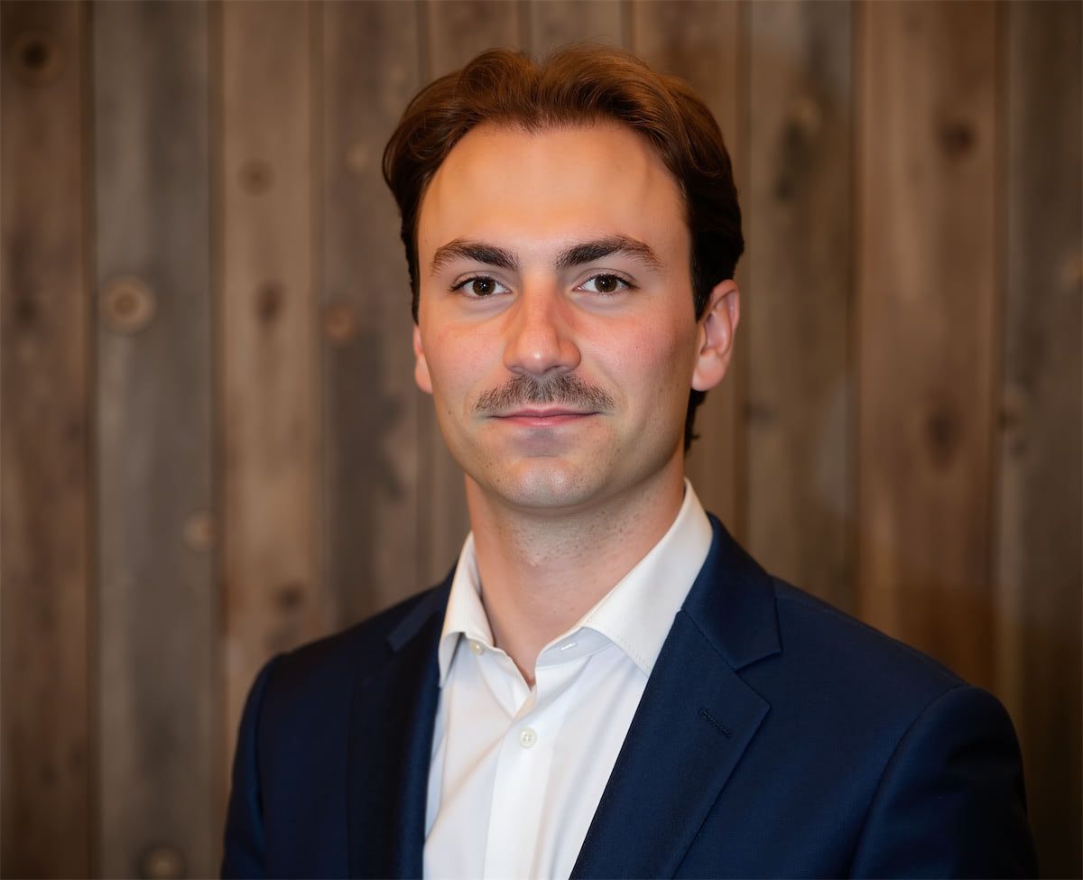 A professional headshot of a person with dark hair and a light mustache, wearing a white shirt and dark blue blazer.