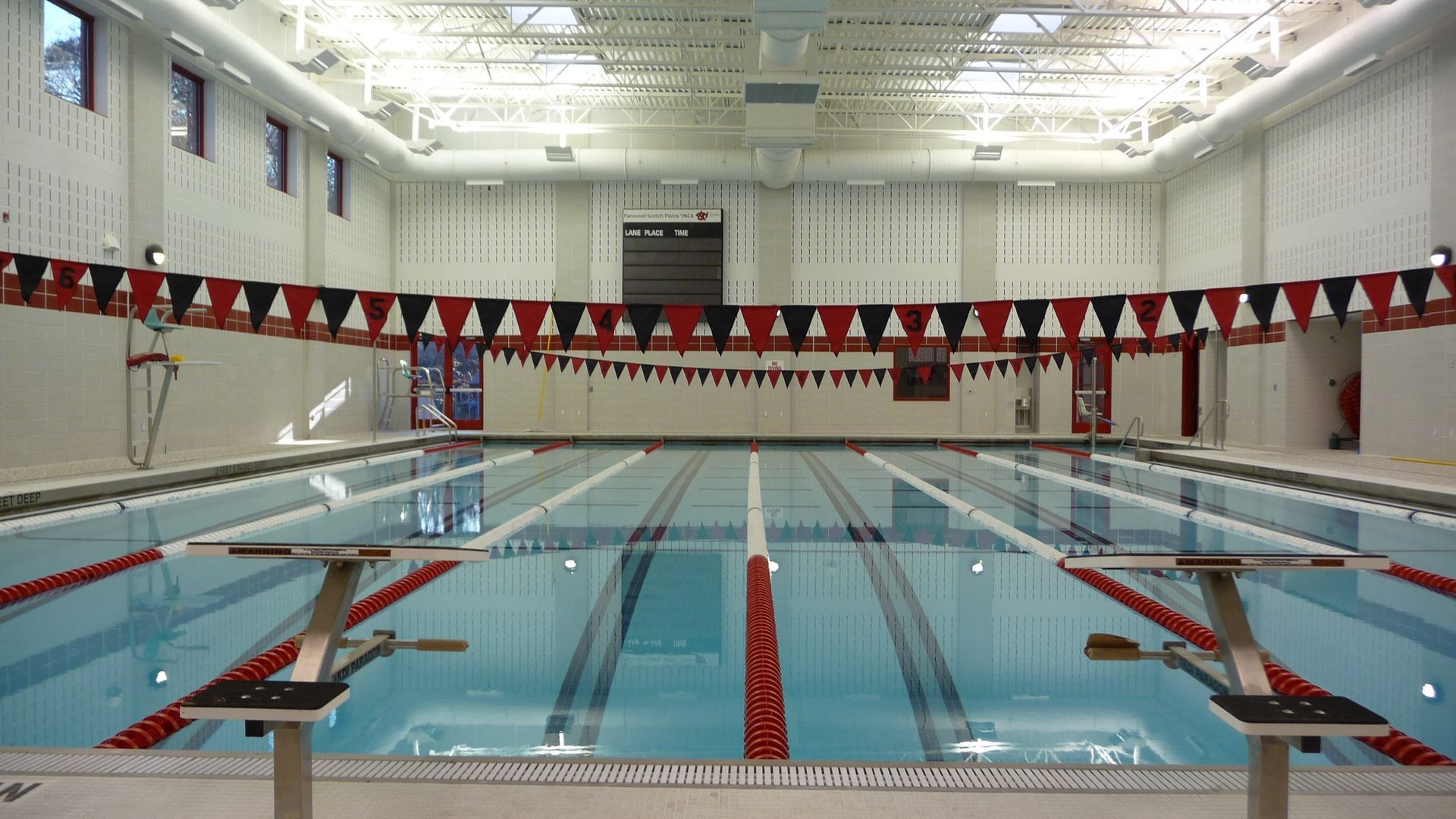 Indoor swimming pool with lanes, starting blocks, and red and black pennant flags hanging above the water.