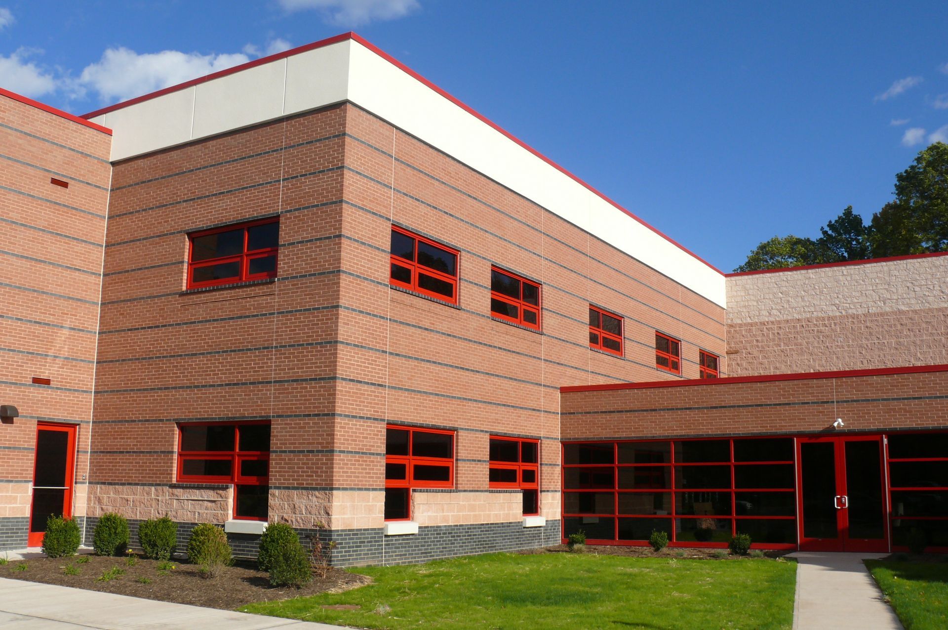 Modern two-story brick building with red-framed windows and doors under a blue sky with clouds.