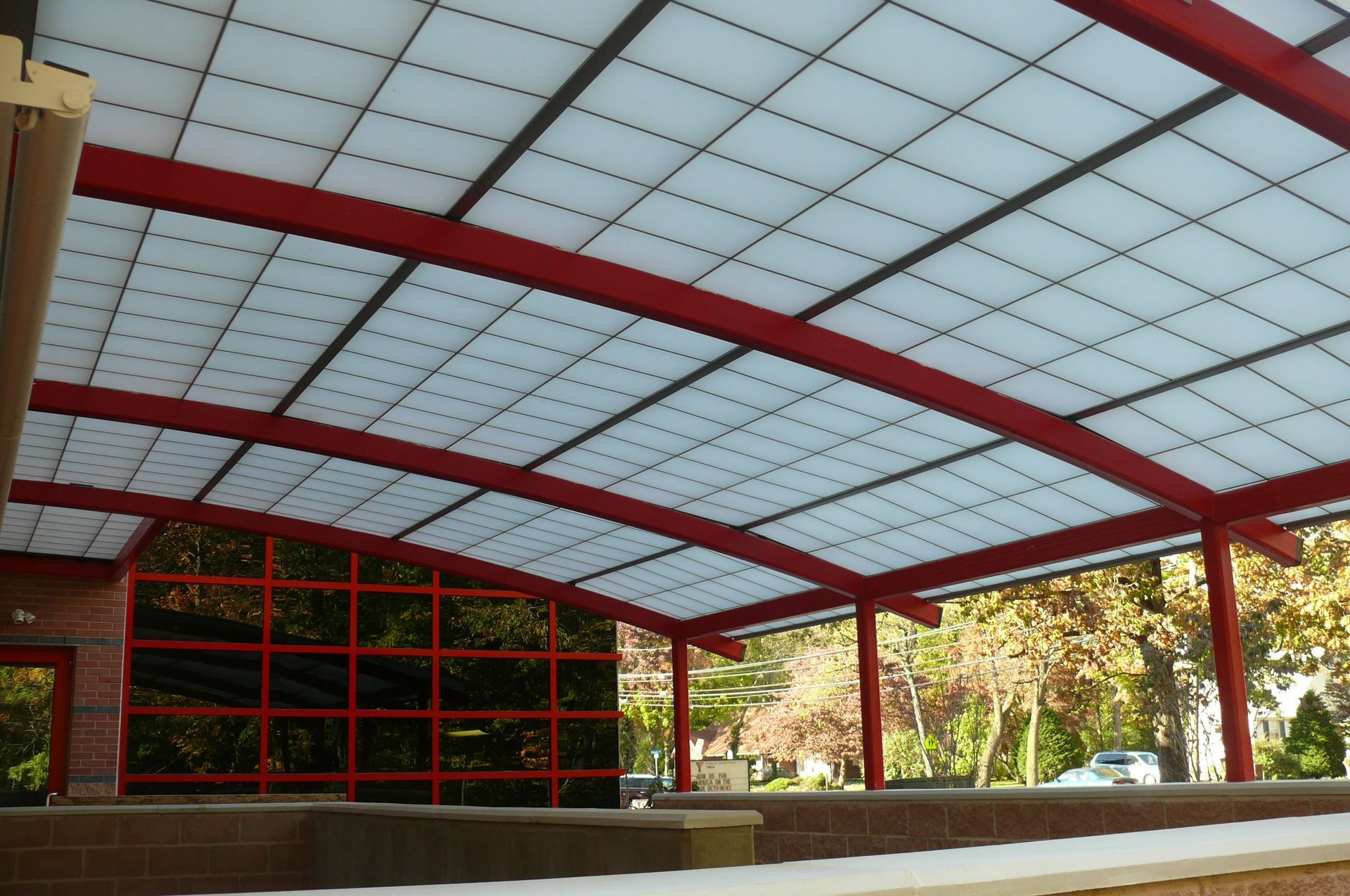A curved, translucent panel canopy supported by a red steel frame, adjacent to a building with red grid window frames.