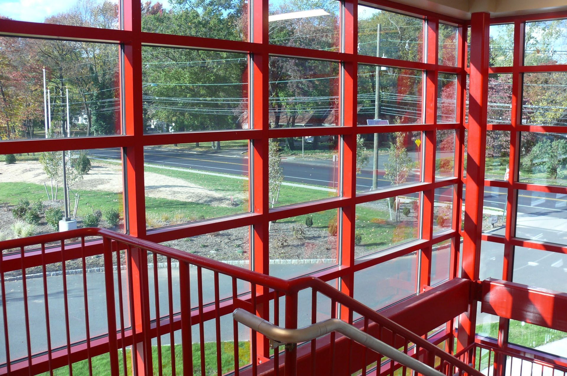 A stairwell with red railings and a large glass window wall overlooking a road, trees, and landscaping.