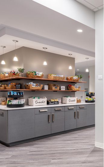 A break room with gray cabinets, white counters, and wooden shelves holding organized baskets of snacks.