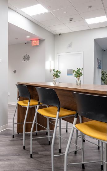 A row of three stools with yellow seats and black backs set against a long wooden counter in a modern, light-filled office.