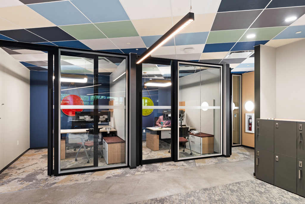 Modern office interior with two glass-walled private workspaces, a multi-colored tile ceiling, and a grey carpet floor.