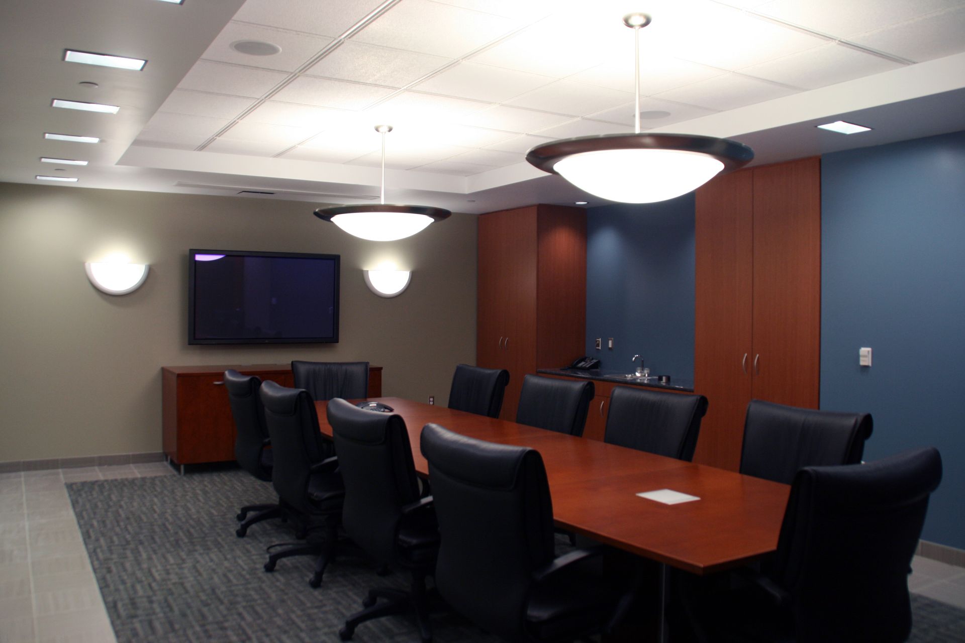 Modern meeting room featuring a long conference table, white chairs, glass walls, and a large display screen on the wall.