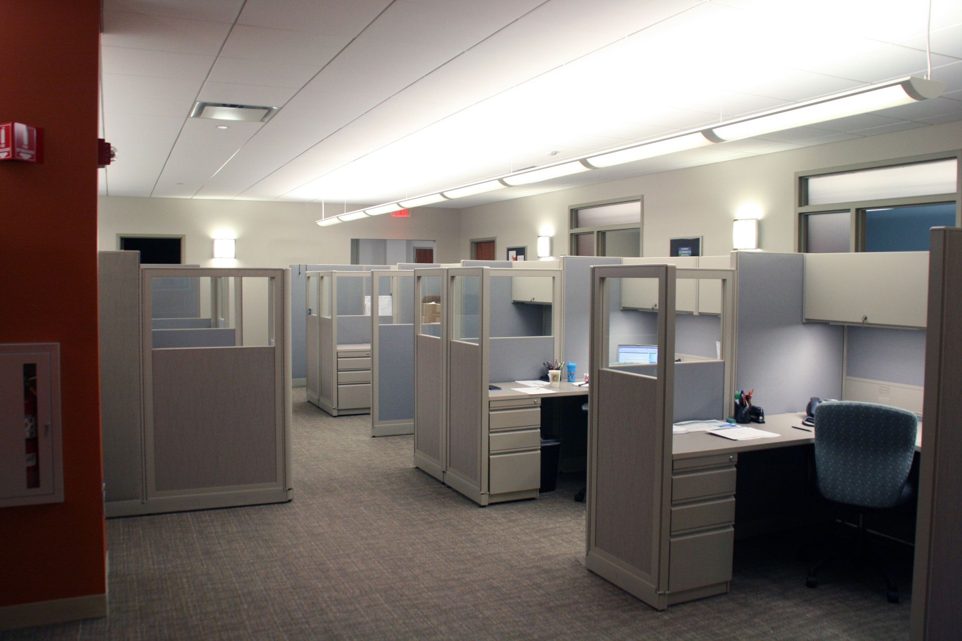 A modern office cubicle with a desk and chair next to a glass wall looking into a warehouse with yellow guardrails.