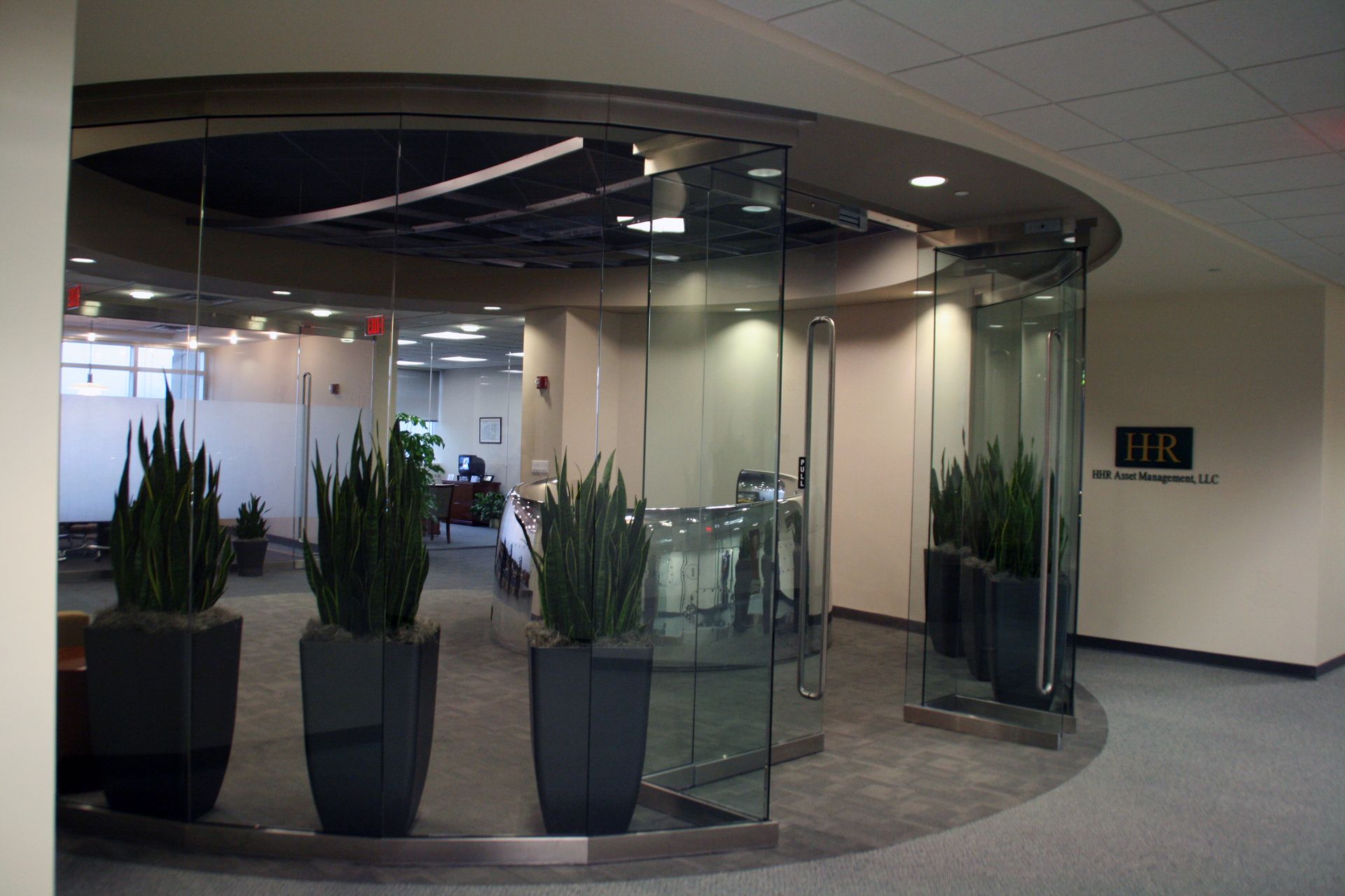 An office entrance with a curved glass wall, three large potted plants in the foreground, and a sign labeled 