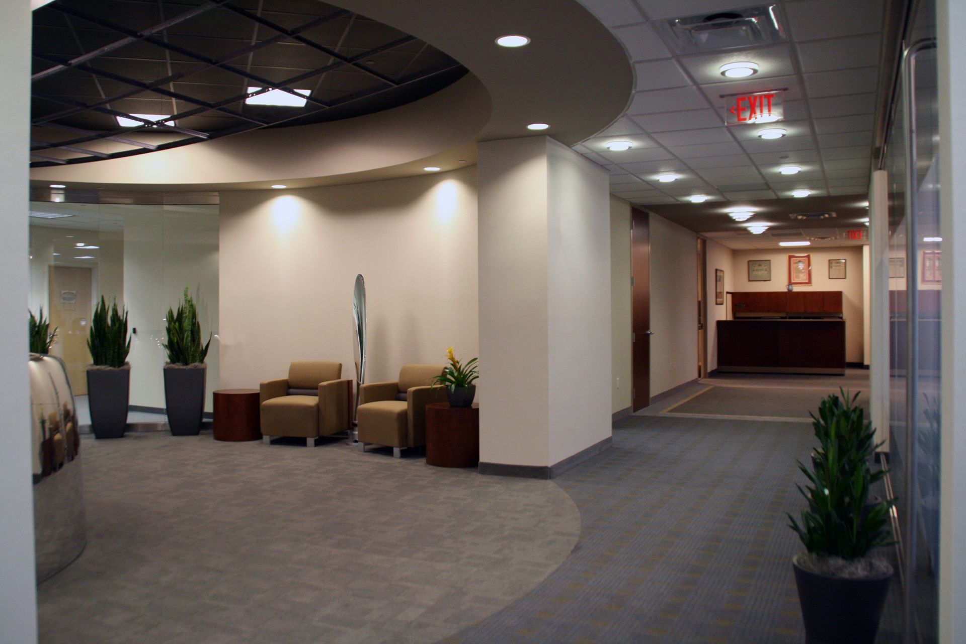 A modern office lobby with a curved ceiling, two brown armchairs, potted plants, and a hallway leading to a reception desk.