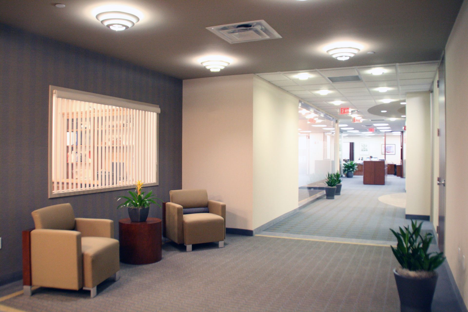 A waiting area with two beige armchairs, a round wooden table with a plant, and a hallway leading into an office space.