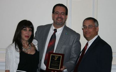 Three people pose for a photo as the center person holds a YMCA award plaque against a plain white wall background.