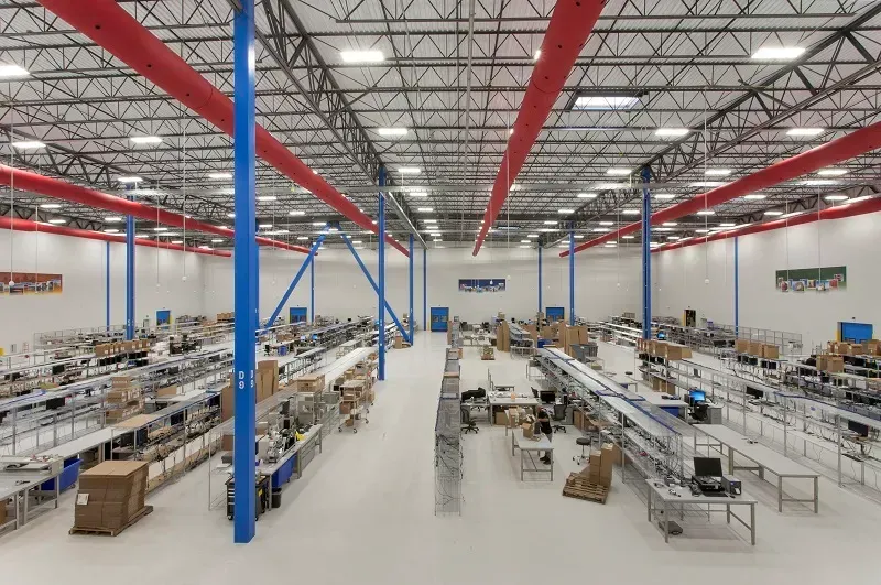 Wide-angle view of a large, high-ceiling warehouse interior with rows of work tables, metal shelving, and red ducting.