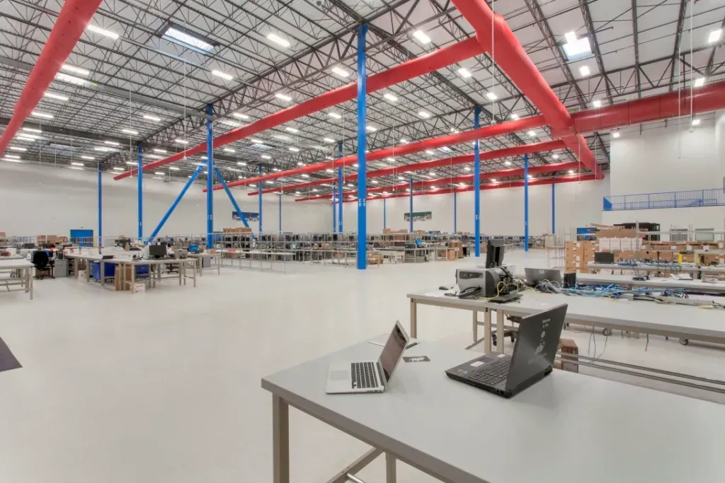 A spacious, well-lit warehouse office with tables, chairs, and laptops, featuring high ceilings and bright red beams.