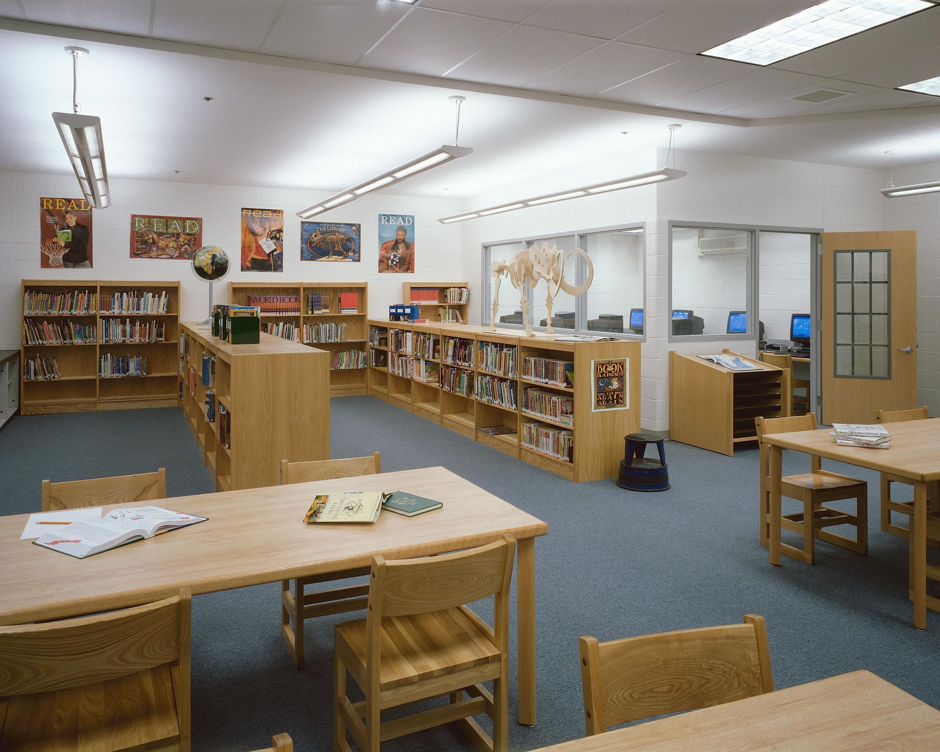A school library with rows of wooden bookshelves, study tables, chairs, and a glass-walled computer lab in the background.