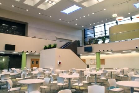 A bright, modern cafeteria with rows of white round tables and chairs under a high ceiling with recessed lighting.