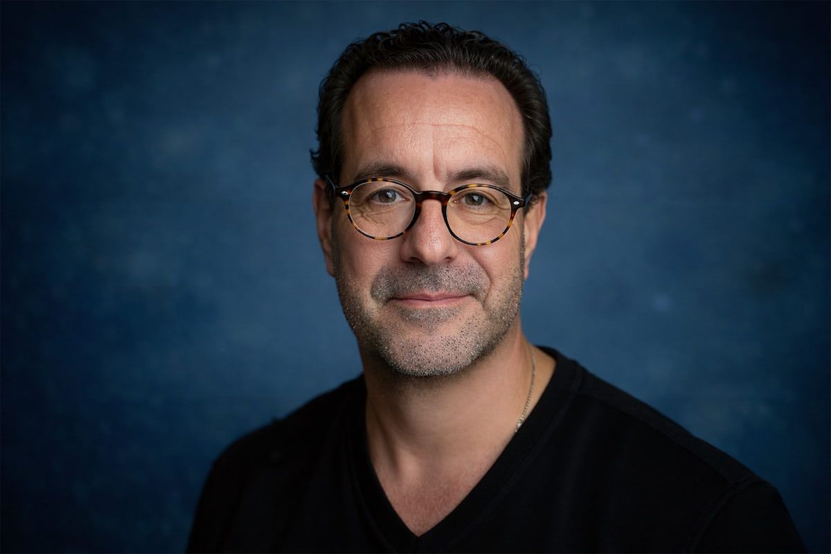 A person wearing glasses and a black shirt smiles slightly against a textured blue background.