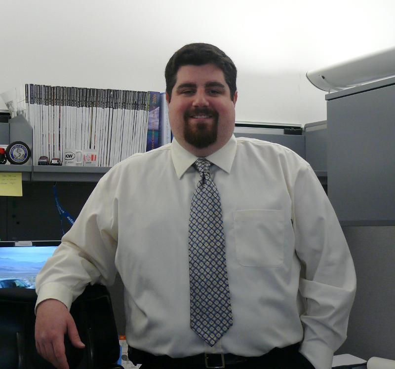 A person with dark hair and a goatee smiles in an office, wearing a white button-down shirt and a patterned gray tie.