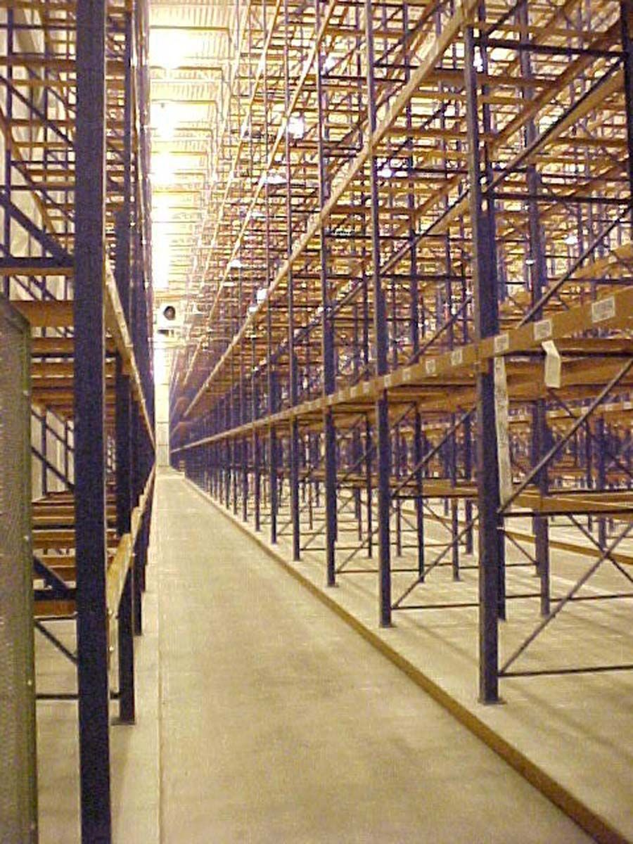 Empty warehouse with rows of tall, industrial metal storage shelving flanking a central concrete aisle.