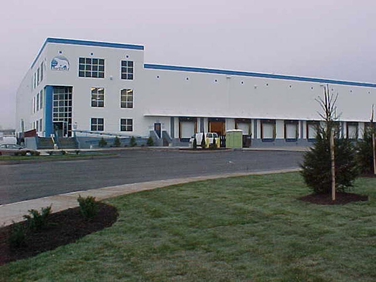 A multi-story, white industrial warehouse with a blue roof, glass entryway, and numerous loading bays.