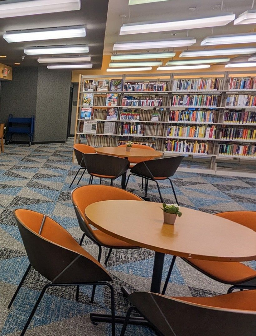 A library seating area with orange chairs and wooden tables on a patterned carpet in front of rows of bookshelves.