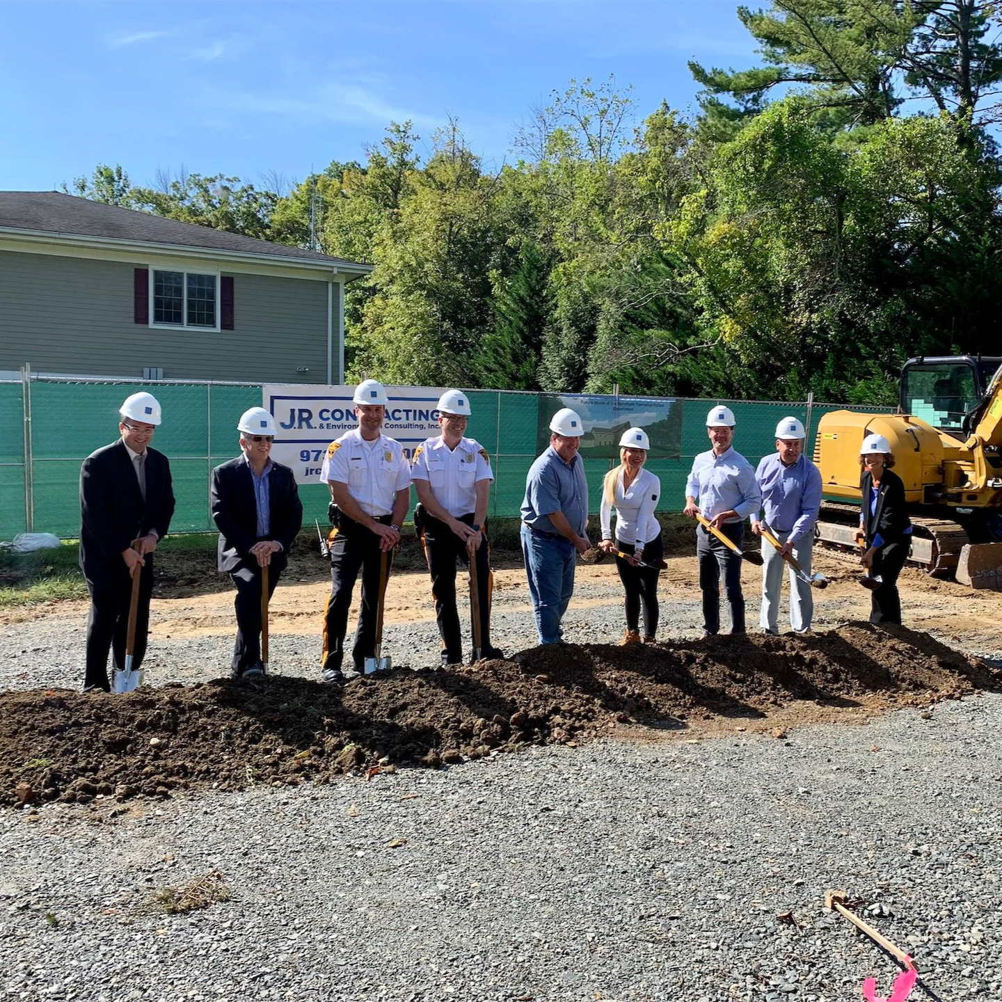 A group of people wearing hard hats perform a ceremonial groundbreaking with shovels at a construction site.
