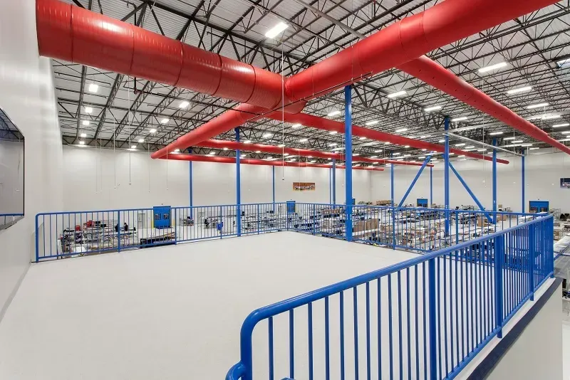 A mezzanine platform with blue railings overlooks a vast warehouse featuring red industrial ductwork on the ceiling.