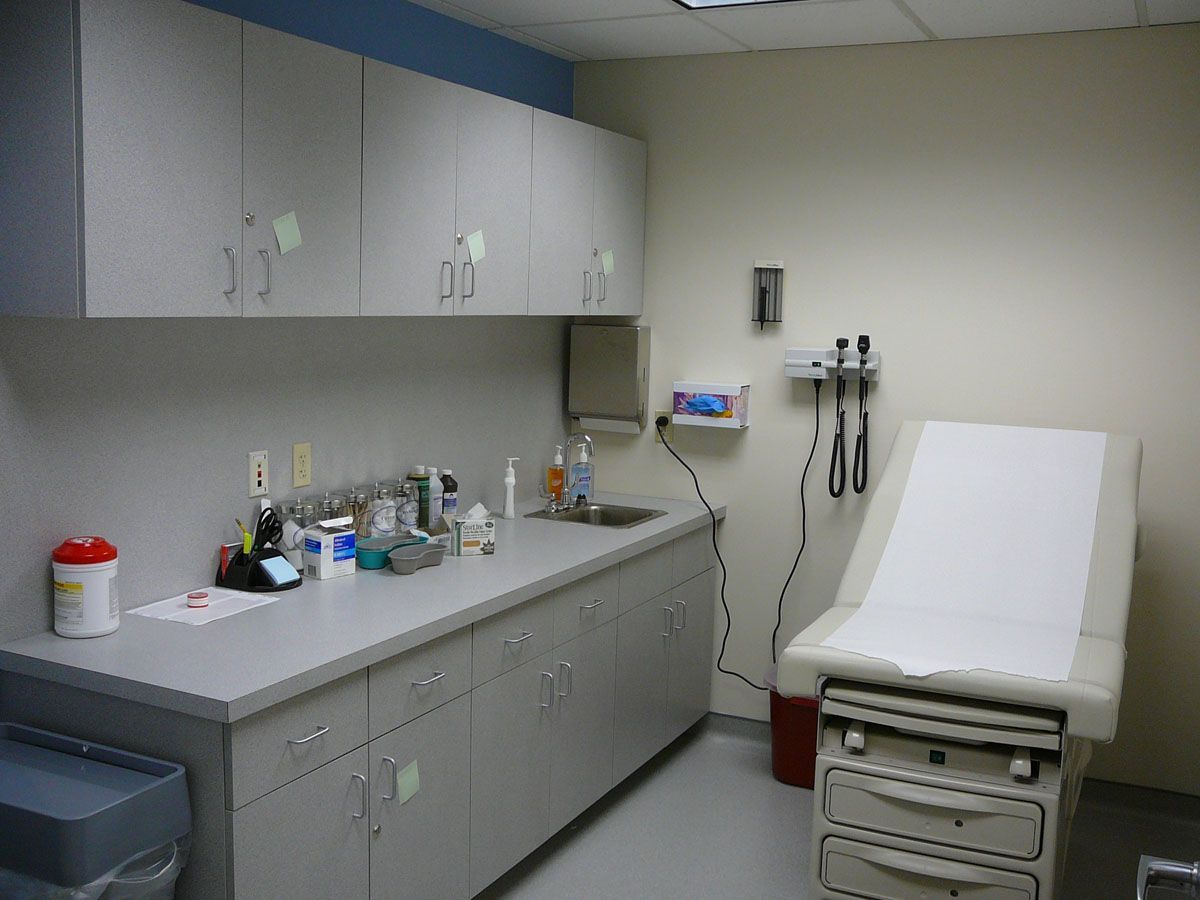 A doctor's examination room with a grey counter, cabinets, a sink, and an exam table with white paper.