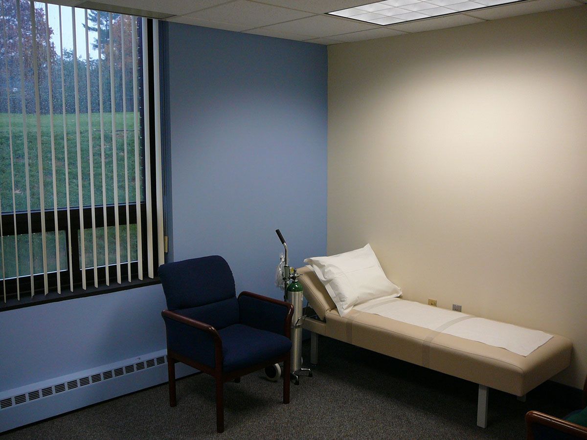 A small medical exam room with a beige treatment table, a navy blue armchair, and a window with vertical blinds.