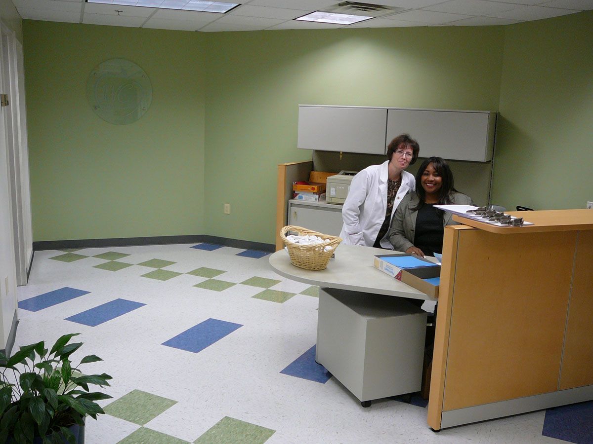 Two office staff members smiling behind a reception desk in a green-walled room with patterned tile floors.