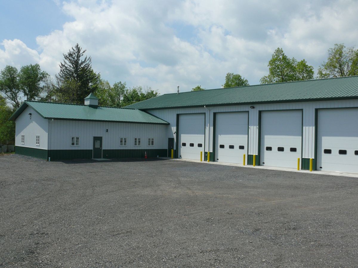 A gray metal maintenance building with a green roof, four white garage doors, and a gravel parking lot.