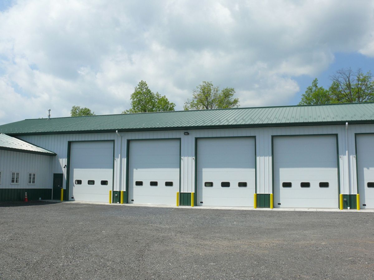 A long commercial building with a green metal roof, light gray walls, and several white garage doors on a gravel lot.