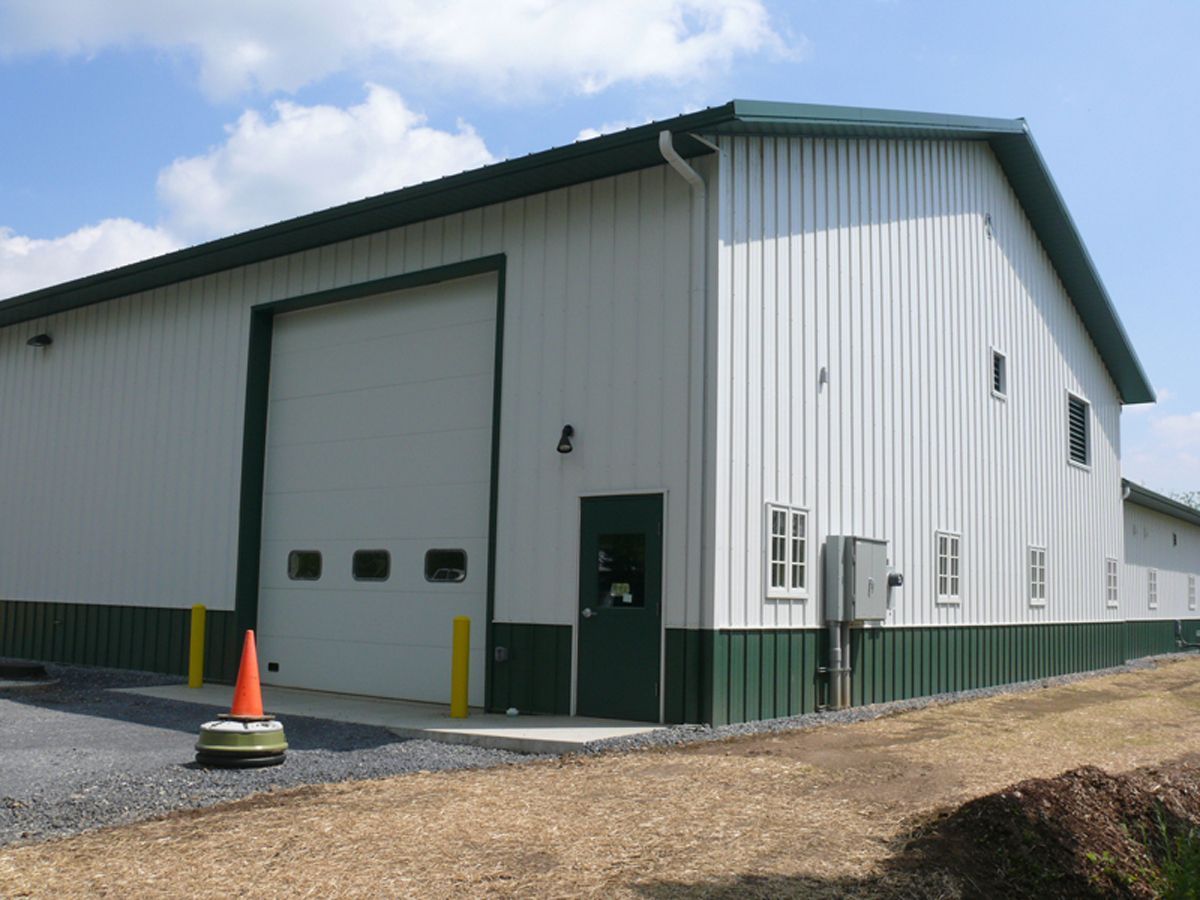 A white and dark green metal warehouse building with a large garage door, a single door, and windows.