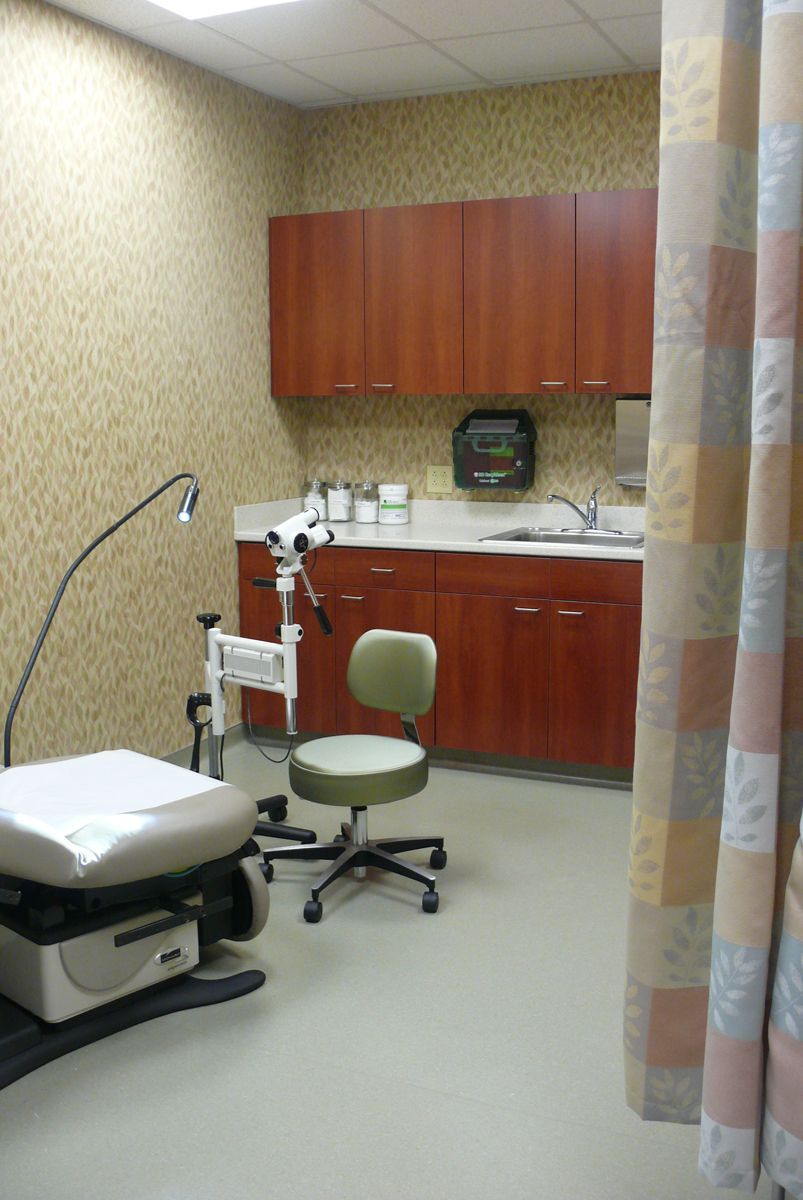 A medical exam room featuring a paper-covered exam table, a rolling stool, a colposcope, and wood-toned cabinetry.