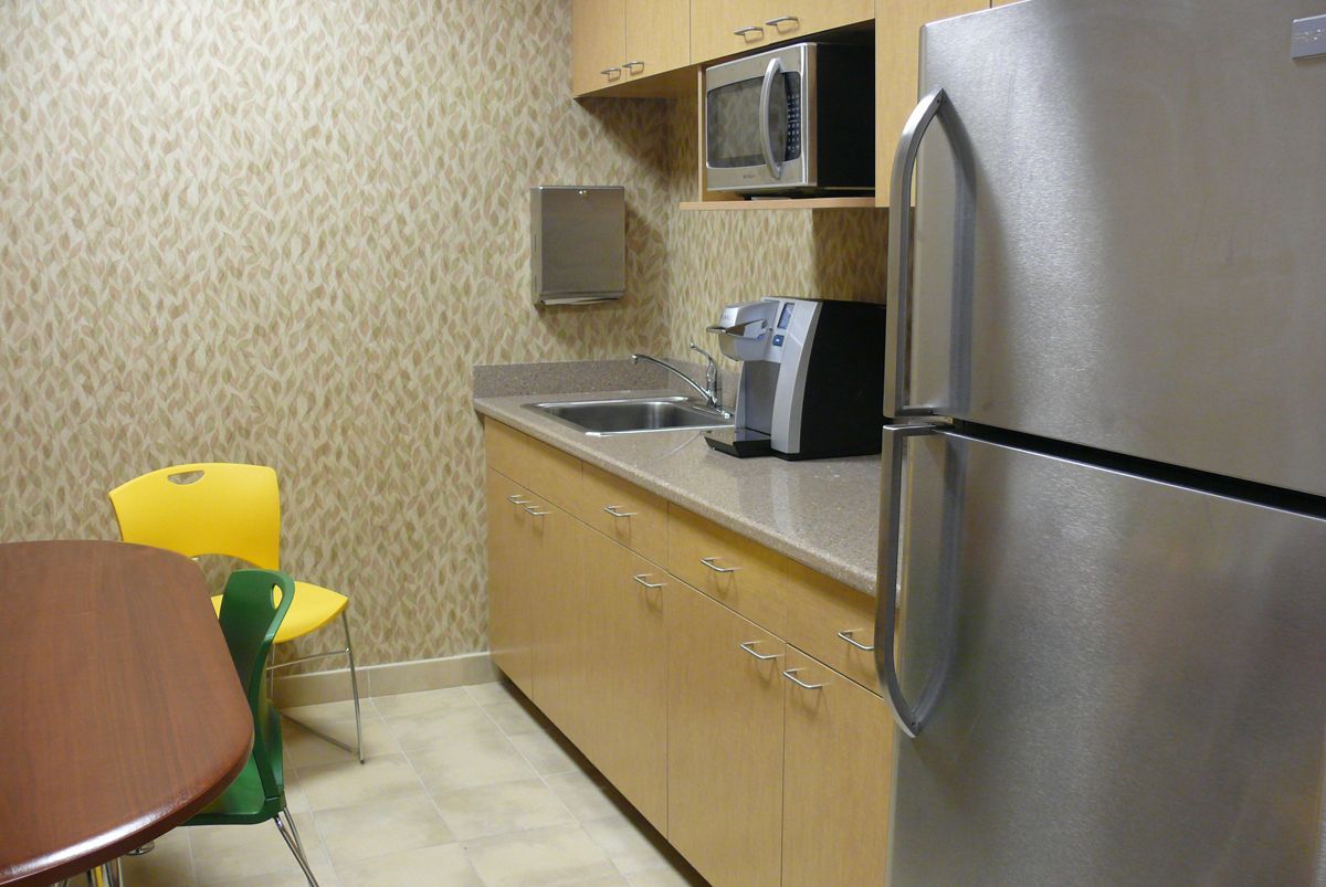 A small breakroom with wooden cabinets, a stainless steel refrigerator, microwave, coffee maker, and chairs at a table.