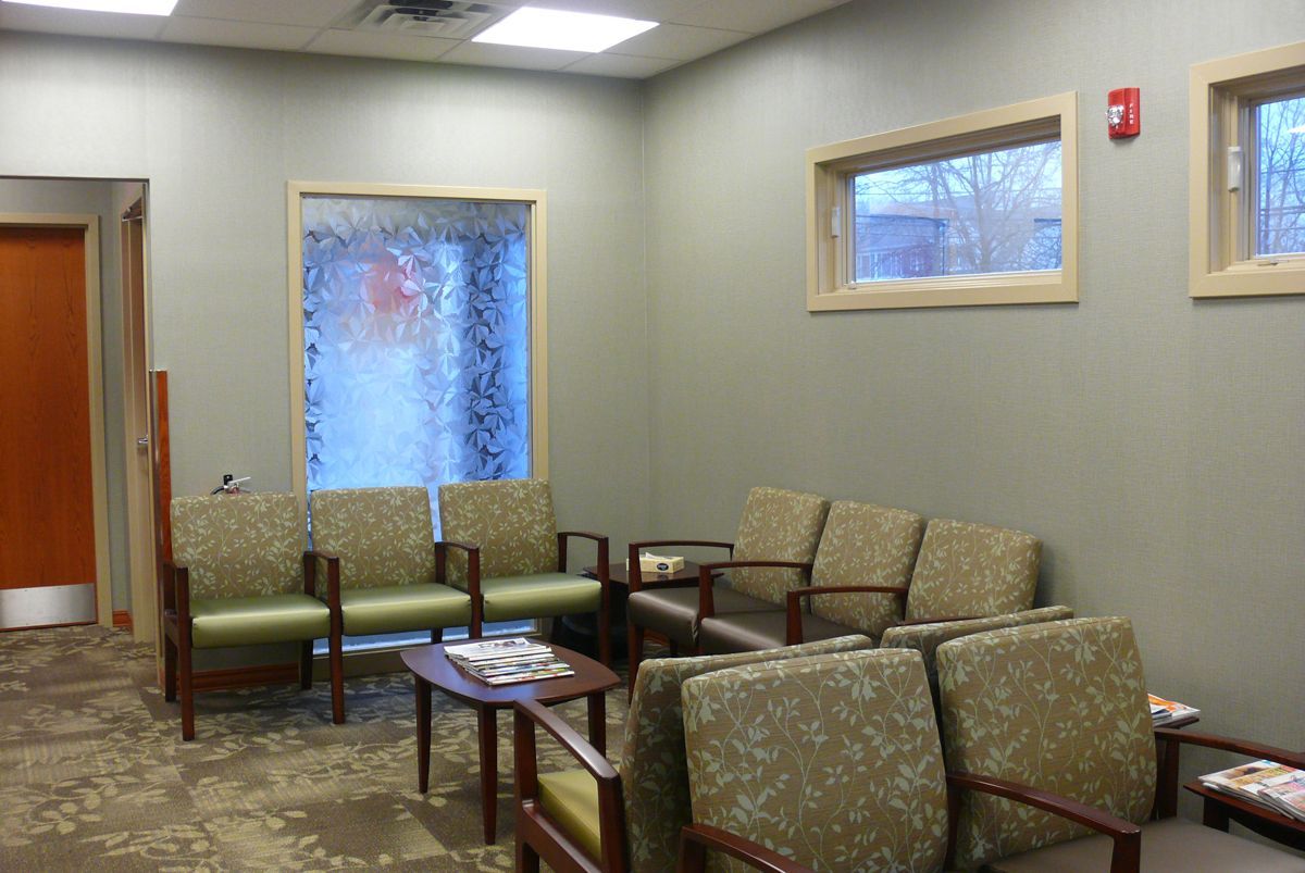 Waiting room with patterned chairs, a small table, a frosted glass window, and two high rectangular windows on beige walls.