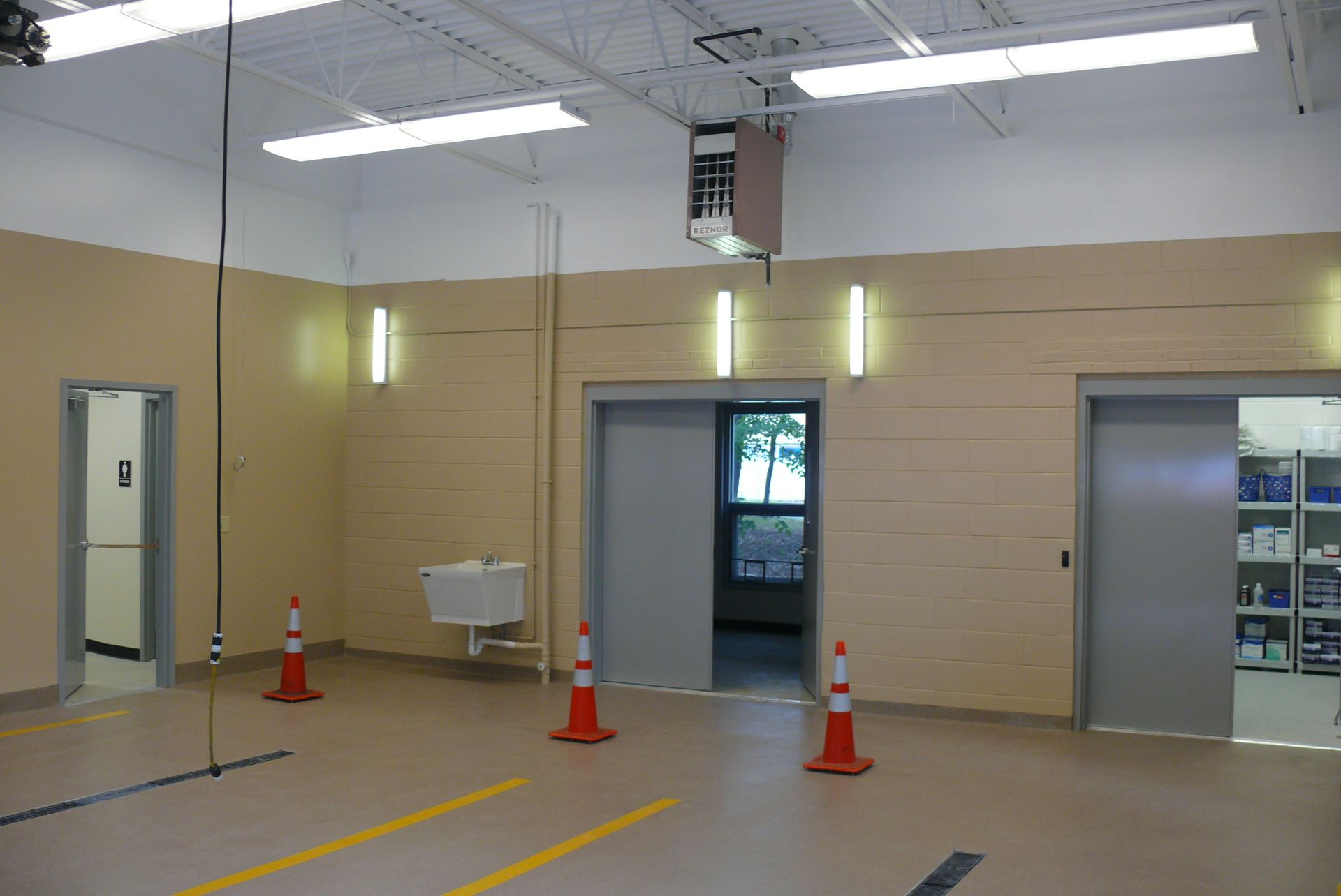 An indoor hallway with beige walls, three doorways, yellow floor markings, and traffic cones.
